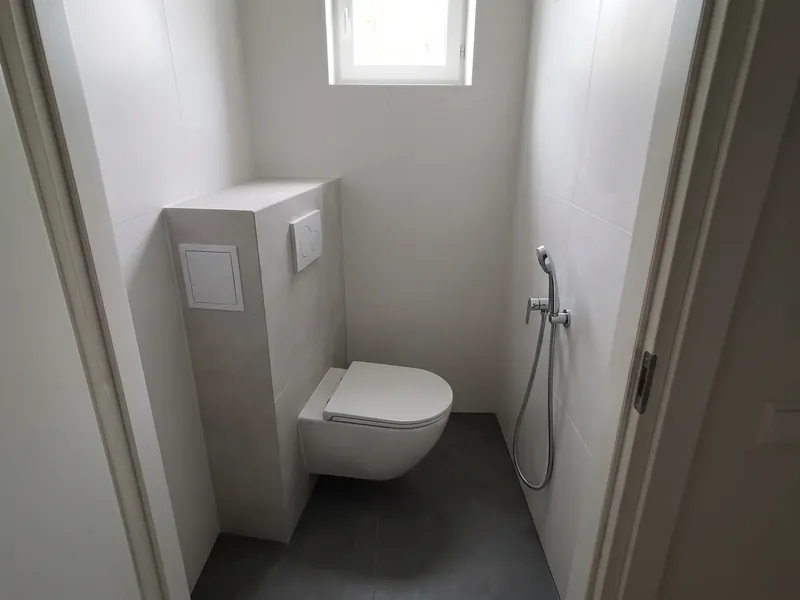 Kitsas WC-ruum seinapoti, plaaditud raamikatte, valge ruudukujulise luugi ja hügieenidušiga, all tumedad põrandaplaadid, seina-WC pott riputatakse nii, et istme kõrgus põrandast on 42 sentimeetrit.